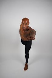 Shirtless tattooed athlete balancing on one leg in a Warrior III yoga pose, reaching forward in black leggings against a clean white studio background.