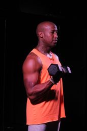 Athletic man in a bright orange tank top performing a dumbbell bicep curl in a dim indoor gym, beaded bracelet and neck tattoo visible.