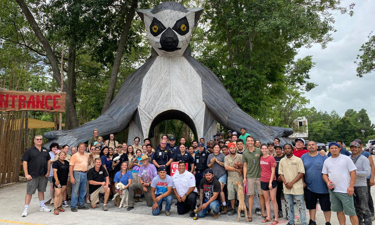 Playful giant wooden lemur sculpture with outstretched arms forming a tunnel at a forested park entrance, a large mixed-age group posing for a daytime group photo beneath the structure next to a red “Entrance” sign.