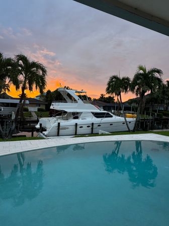 Sunset over a tropical canal with a white luxury yacht docked by a waterfront home, palm trees and a blue swimming pool reflecting the sky.