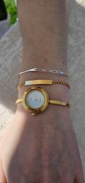 Sunlit close-up of a wrist wearing a round gold bangle watch and two stacked bracelets — a delicate gold chain with an engraved nameplate and a thin silver chain — over light skin and a textured beige background.