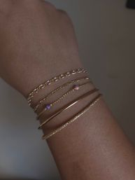 Close-up of a wrist wearing a stack of delicate gold bracelets — twisted chain, smooth bangle, pearl-linked chain and tiny opal bead accents on a neutral background