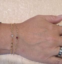 Close-up of a wrist wearing two delicate gold chain bracelets with small bezel-set stones (clear and red) and a silver ring, photographed on a pale satin background.