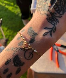 Close-up of a tattooed forearm wearing a gold heart charm bracelet with tiny pearls, sunlit backyard grass and red-handled pliers blurred in the background.