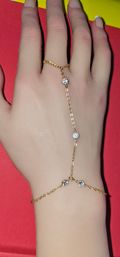 Dainty gold-tone hand chain bracelet connecting a ring to a wrist with three sparkling bezel-set clear stones, shown on a hand over a red background