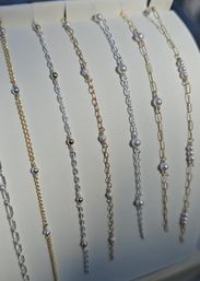 Close-up of delicate gold and silver chain bracelets with tiny pearls and metallic bead accents displayed vertically on a white jewelry stand — minimalist fashion accessory close-up