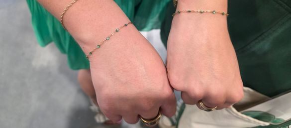 Close-up of two hands side-by-side wearing delicate gold chain bracelets with small green stones and matching gold rings, green dress visible in the background — fashion jewelry detail.