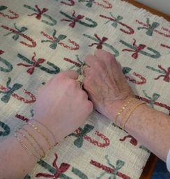 Two hands — one younger and one older — resting side-by-side on a festive Christmas candy-cane patterned tablecloth, both wearing gold bracelets and rings, cozy holiday scene.