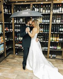 Bride and groom kiss under a black umbrella in a wine shop aisle, groom in a tuxedo and bride in a white gown surrounded by wooden shelves stocked with bottled wine — romantic indoor wedding photo.