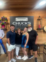 Four smiling adults posing arm-in-arm inside a wood-paneled wine and spirits tasting room in front of a large black sign, with festive Mardi Gras beads and lantern decor.