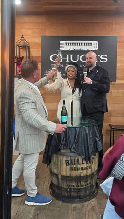 Three people raising champagne flutes in a celebratory toast at a rustic indoor tasting room, glass-top barrel table with bottle and wood-paneled wall backdrop.