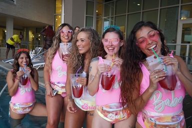 Five women at a poolside bachelorette-style party in matching pink swimsuits and pastel fanny packs, laughing and sipping red drinks from clear pouch cups while wearing sunglasses.