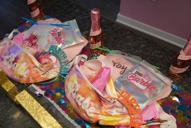 Iridescent "Babe" fanny packs, Barbie-themed pouches, mini rosé bottles, party tiaras and colorful sequin runner arranged as party favors on a kitchen countertop