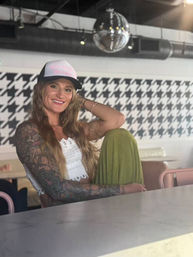 Smiling tattooed woman in a trucker hat and white crochet top seated at a marble counter in a trendy café with houndstooth wall pattern, disco ball, and green skirt.
