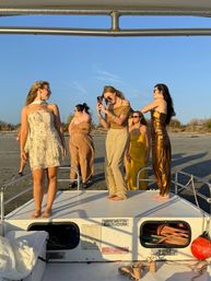 Group of friends in gold and cream summer dresses posing and taking photos on the bow of a boat at golden-hour on a sandy beach