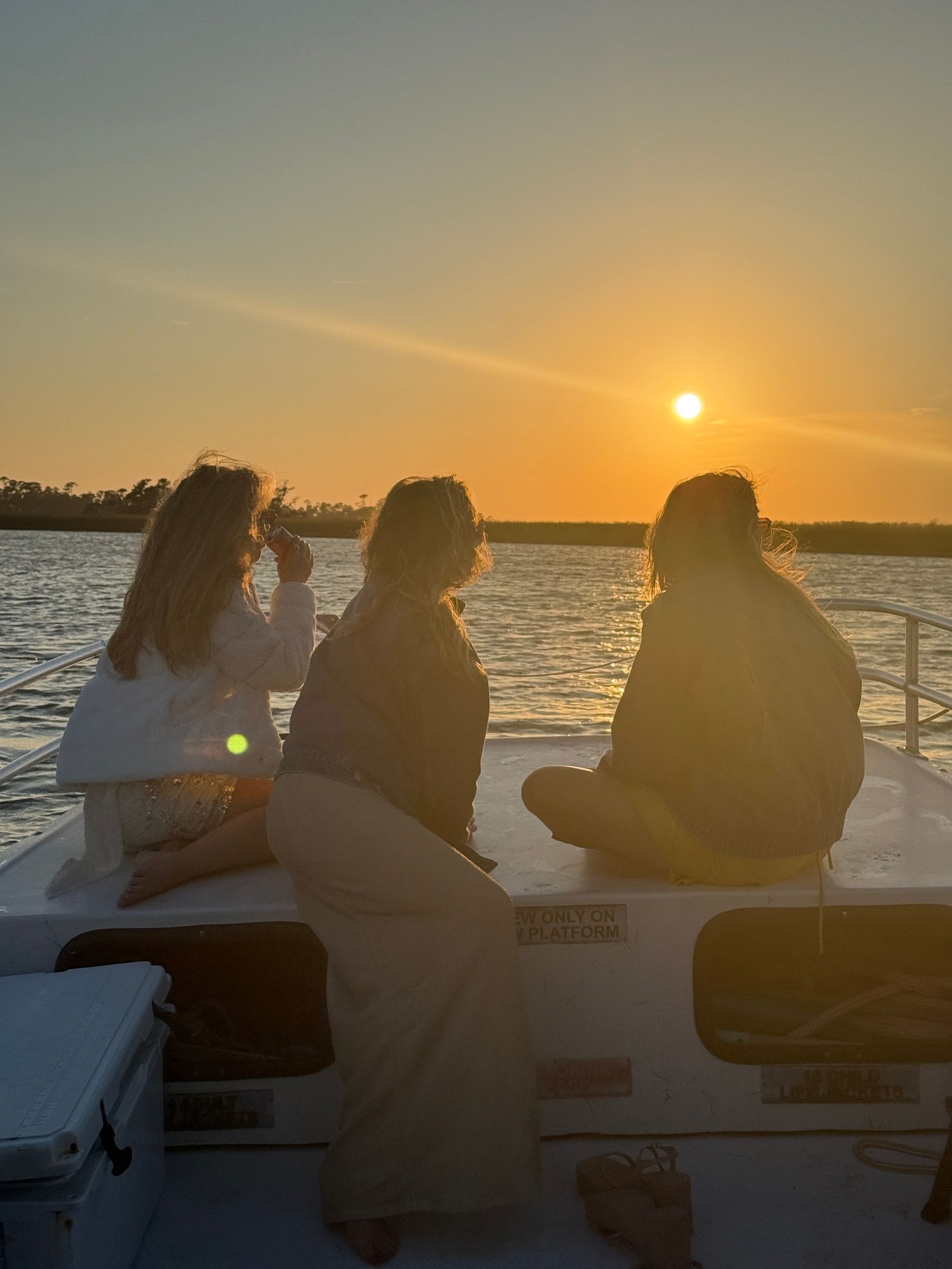Three people sitting on a small boat’s bow watching a golden sunset over calm coastal bay waters during a sunset boat ride
