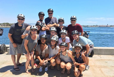 Smiling mixed-age group wearing helmets posing on a sunny waterfront pier beside rental electric scooters with blue bay water and distant shoreline in the background.
