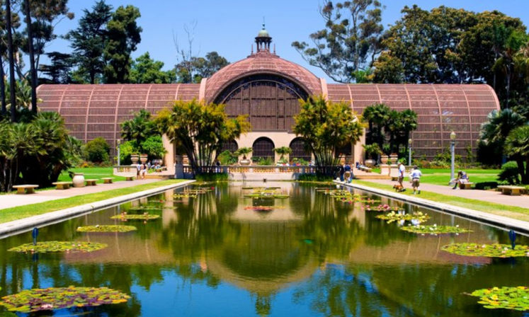 Sunlit arched conservatory mirrored in a lily-pad pond, framed by palm trees and visitors strolling along manicured garden paths.