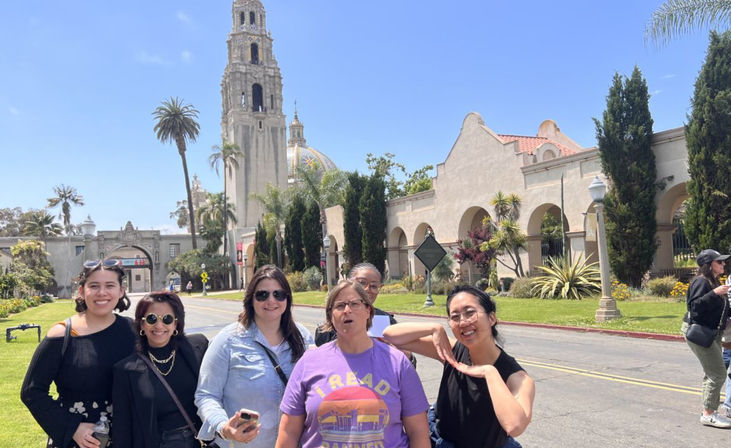 Six adults smiling and posing under blue skies with palm trees in front of the Spanish-style California Tower at Balboa Park, San Diego.