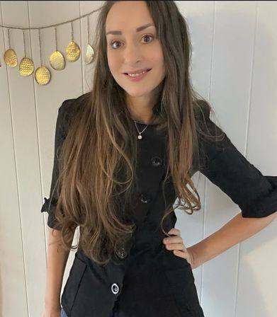 Smiling woman with long brown hair wearing a black buttoned jacket and pendant necklace, posing hand on hip against a white paneled wall with hanging gold disc decor — casual indoor portrait.