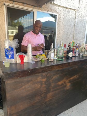 Smiling man in a pink polo mixing drinks at an outdoor backyard patio home bar with liquor bottles, limes, plastic cups and cocktail tools