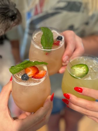 Three colorful summer cocktails clinking in stemless glasses, two pink berry-mint drinks garnished with strawberries, blueberries and mint, and one green jalapeño-lime cocktail, hands with red nails toasting.