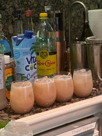 Four frosty pink coconut cocktails in stemless glasses on a granite kitchen counter with Vita Coco carton, Topo Chico bottle, cocktail shaker and bar tools — home bar setup.