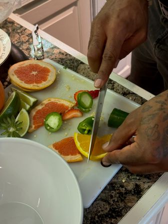 Tattooed hands slicing an orange on a white cutting board in a home kitchen, with grapefruit halves, lime wedges, sliced jalapeño and strawberries on a granite countertop next to a white bowl.