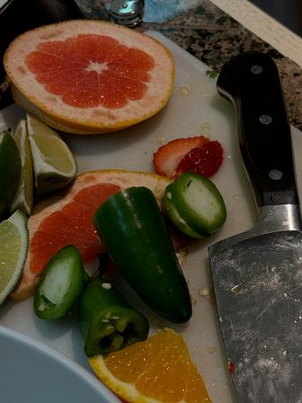 Vibrant kitchen cutting board on granite countertop with halved pink grapefruit, lime wedges, sliced jalapeños, an orange wedge, a strawberry slice and a cleaver-style knife