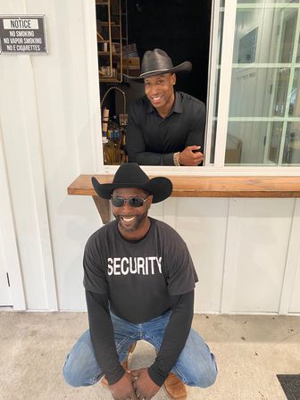 Smiling duo in cowboy hats at a white service window — one kneeling in a 'SECURITY' shirt and sunglasses, the other leaning on the counter in a black shirt.