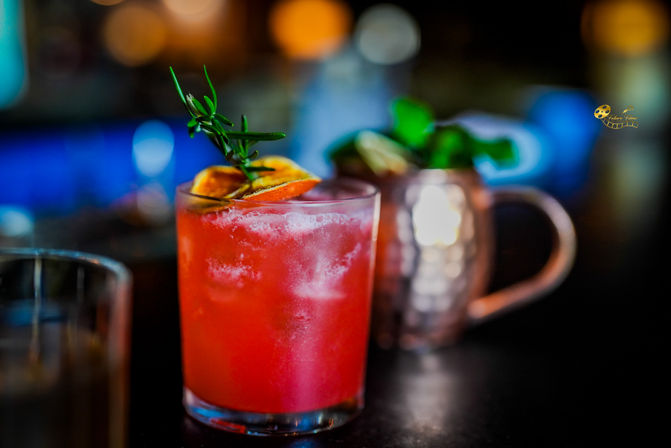 Vibrant red cocktail in a short glass with crushed ice, garnished with a dried orange slice and rosemary sprig, with a blurred copper mug and colorful bokeh bar lights in the background.