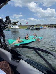 Four adults lounging on a bright turquoise floating mat near anchored boats on a sunny lake, blue sky with fluffy clouds and a grassy shoreline in the background.