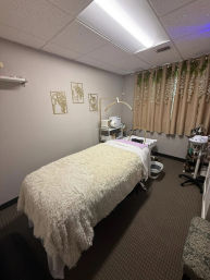 Cozy spa treatment room with plush faux-fur treatment table, neutral curtains with hanging greenery, gold arched lamp and skincare equipment cart.