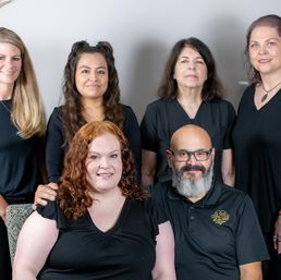 Smiling diverse team portrait of six adults in black shirts, mixed ages and genders, posed indoors against a neutral background — casual professional staff photo for a small business.