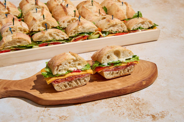Bite-size ciabatta sandwiches with salami, cheddar, lettuce and mayo on a wooden board, with a full catering platter of sliders in the background