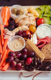 Colorful charcuterie box: grapes, carrot sticks, crackers, sliced bread, cherry tomatoes, cucumber ribbons, blackberries and dried cranberries, hummus and creamy dip, prosciutto and salami, dark chocolate pieces and cookies — picnic-ready grazing platter.