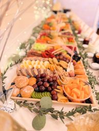 Elegant grazing table with wooden charcuterie boards piled with grapes, kiwi, melon cubes, sliced meats, cheeses, pastries and powdered donuts, garnished with eucalyptus and twinkling fairy lights for a festive event spread.
