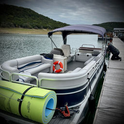 Pontoon boat docked at a wooden lake pier under an overcast sky, life ring and rolled green foam mats on deck, person kneeling to secure a rope.