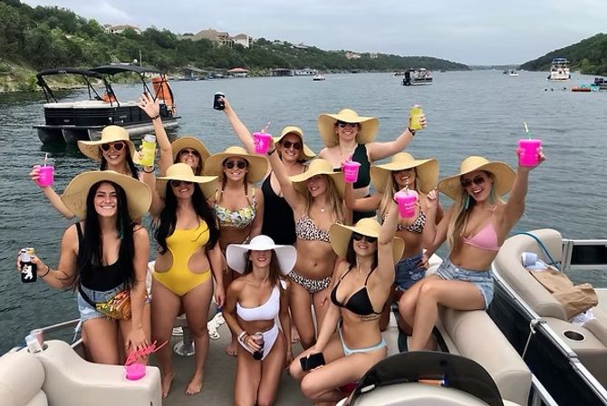 Group of women in wide-brim sun hats and swimsuits cheering with neon cups on a pontoon boat at a scenic lake shoreline