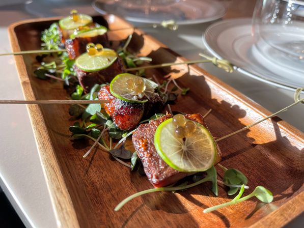 Skewered glazed pork belly bites topped with thin lime slices and citrus pearls, served on microgreens on a wooden rectangular platter in bright natural light