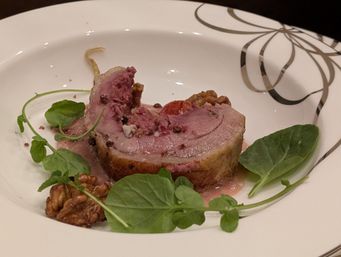 Plated rolled roast lamb with pink jus, cracked peppercorns, watercress and candied walnuts on a white decorative dinner plate — elegant fine-dining presentation.