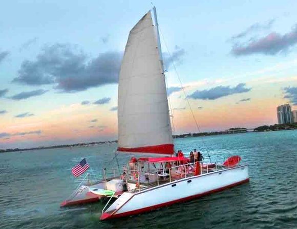 White-and-red catamaran sailboat with tall sail and American flag gliding on turquoise coastal waters at sunset with people on deck and distant skyline