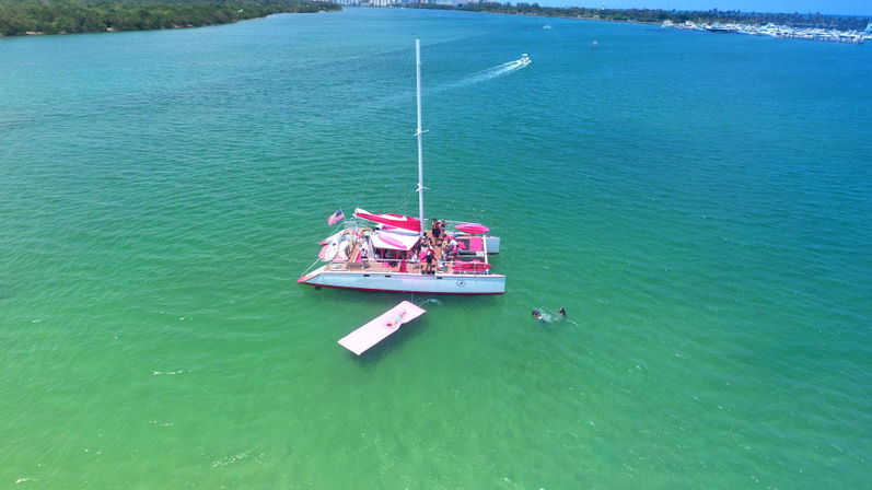 Aerial view of a pink-and-white party catamaran anchored in clear turquoise water with a pink floating platform, people on deck, two swimmers nearby and a marina-lined coastline in the distance.
