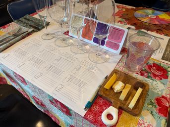 Wine tasting table setup with four empty wine glasses on a printed tasting sheet, a small wooden board with four cheese samples and a piece of chocolate, a pen, patterned plastic water cup, and colorful floral tablecloth.