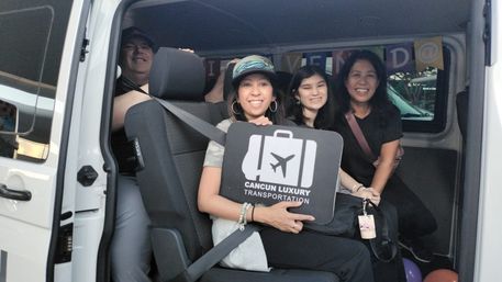 Four smiling travelers inside a white shuttle van holding a luggage sign with an airplane icon, posing for a vacation airport transfer photo.