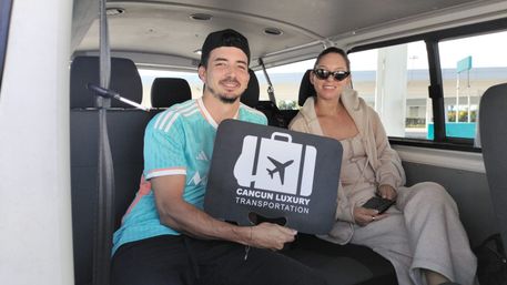 Two smiling travelers seated in a Cancun shuttle van holding a black suitcase-shaped airport transfer sign with an airplane icon