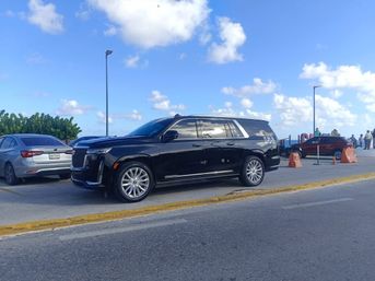 Shiny black luxury SUV parked in an oceanfront parking area under a bright blue sky, with other cars, orange traffic cones, lamp posts and people on a coastal promenade.