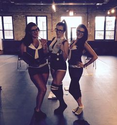 Three women in matching pinstripe costume outfits and glasses pose for a mirror selfie in an industrial loft studio with exposed brick windows, knee-high socks and checkered slippers.