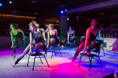 Five dancers in black leotards performing a high-energy chair dance on a nightclub stage lit by vibrant purple, blue and pink spotlights.