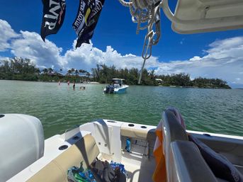From a motorboat deck, view of a shallow turquoise bay with an anchored skiff and people wading near a palm-lined sandy shore under bright blue sky and puffy clouds, ropes and boat gear in the foreground.